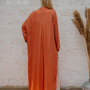 May include: A long, orange, button-down dress. The dress has long sleeves and a relaxed fit. The model is standing in front of a white brick wall. The brand name "chintamani" is printed on the wall.