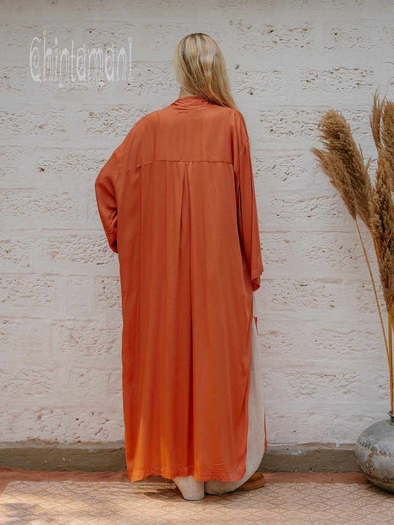 May include: A long, orange, button-down dress. The dress has long sleeves and a relaxed fit. The model is standing in front of a white brick wall. The brand name "chintamani" is printed on the wall.