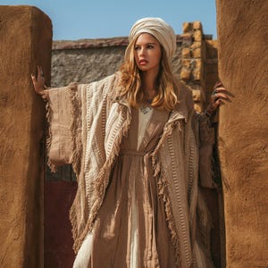 May include: A beige and cream-coloured layered outfit consisting of a long, belted dress and a textured, fringed poncho. The ensemble is completed with a matching beige head wrap. The text Chintamani is visible in the upper left corner of the image.