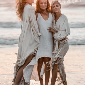 Puede incluir: Tres personas con ropa fluida de color crema de pie en el oc&eacute;ano al atardecer. La imagen incluye el texto "@chintamani alCHEMY". La ropa tiene flecos y detalles drapeados.