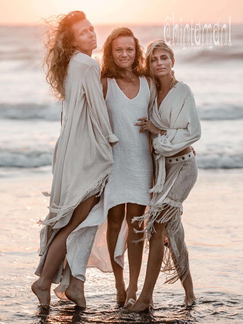 Puede incluir: Tres personas con ropa fluida de color crema de pie en el oc&eacute;ano al atardecer. La imagen incluye el texto "@chintamani alCHEMY". La ropa tiene flecos y detalles drapeados.