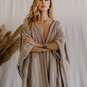 Puede incluir: Un poncho tejido en tonos beige y marr&oacute;n claro con rayas verticales texturizadas y detalles de flecos en los bordes. La prenda tiene un escote en V profundo y mangas anchas, confeccionada con una tela gruesa con un patr&oacute;n de tejido visible.
