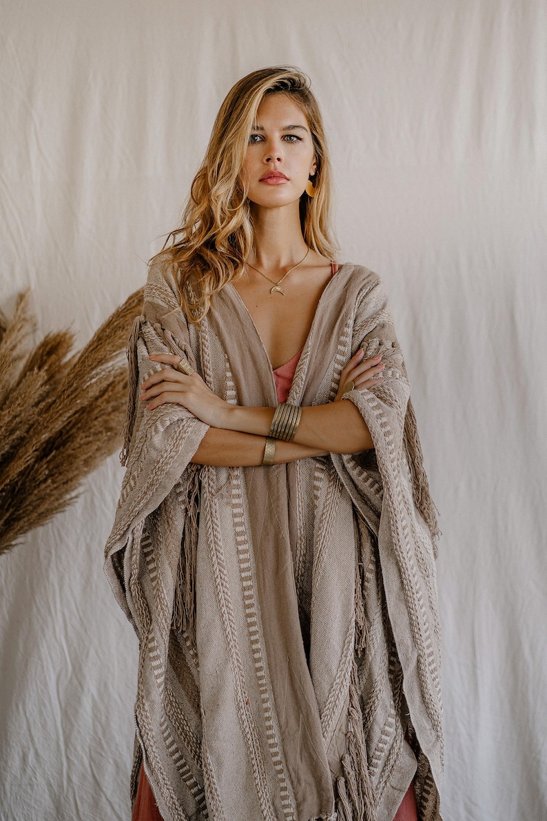 Puede incluir: Un poncho tejido en tonos beige y marr&oacute;n claro con rayas verticales texturizadas y detalles de flecos en los bordes. La prenda tiene un escote en V profundo y mangas anchas, confeccionada con una tela gruesa con un patr&oacute;n de tejido visible.