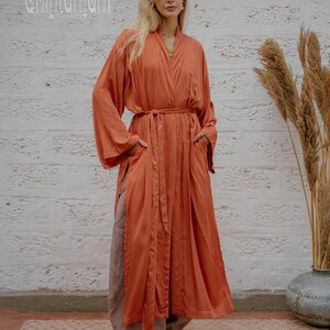 May include: A long, flowing rust-coloured robe with a matching belt and side pockets. The robe is worn over loose, light brown trousers and yellow shoes. The background features a textured white wall and decorative plants. The text "chintamani" is visible in the upper left corner.