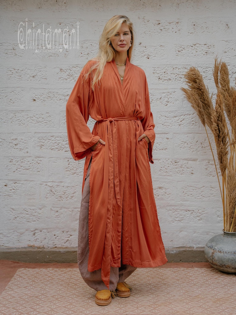 May include: A long, flowing rust-coloured robe with a matching belt and side pockets. The robe is worn over loose, light brown trousers and yellow shoes. The background features a textured white wall and decorative plants. The text "chintamani" is visible in the upper left corner.