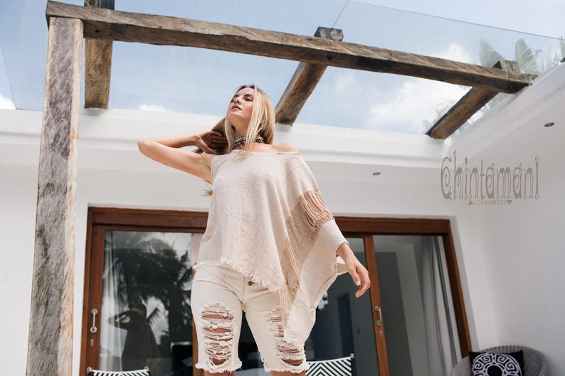 May include: A beige, asymmetrical, sleeveless top with a raw, fringed hemline and a textured, woven fabric finish. The garment features a loose, draped silhouette with a wide neckline and a distinct, long fringe detail along one side.