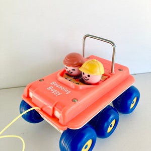 May include: A vintage toy car in coral with blue wheels and yellow accents. The car has two toy figures inside and a metal handle. The text "Bouncing Buggy" is printed on the side, and a yellow string is attached.