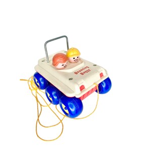 Puede incluir: Un buggy de juguete de plástico blanco con ruedas azules y una franja roja. El buggy tiene un asa y dos figuras en la parte superior. El texto "Bouncing Buggy" está impreso en el lateral.