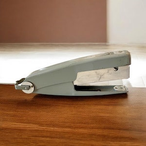May include: A gray stapler with a silver metal staple holder. The stapler is resting on a wooden surface. The text "SOFT CAP TOUCH" is visible on the staple holder.