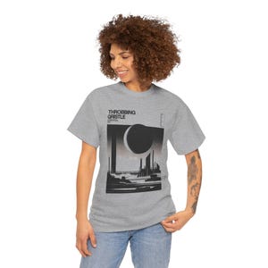 Op de afbeelding: Lichtgrijs t-shirt met de tekst "THROBBING GRISTLE" en een zwart-witte afbeelding van een stadsgezicht met een grote cirkelvorm. Het t-shirt wordt door een persoon gedragen.