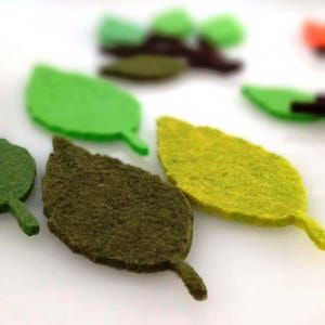 May include: Close-up of felt leaf shapes in shades of green and yellow. The leaves have a textured appearance and are arranged on a white surface. Some leaves are bright green, while others are a darker olive green.
