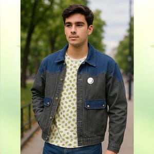 Puede incluir: Chaqueta vaquera de dos tonos, con cuerpo gris oscuro y canesú y cuello azules. La chaqueta tiene dos bolsillos en el pecho y cierre de botones. Debajo, se ve una camiseta blanca con un estampado repetido de aguacates. Un pin plateado está sujeto a la chaqueta.