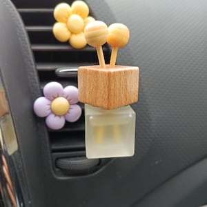 Op de afbeelding: Auto-geurverspreider met een houten blok en twee bolvormige toppen, bovenop een heldere, vierkante fles. Twee bloemvormige ventilatieclips in geel en paars zijn ook zichtbaar.