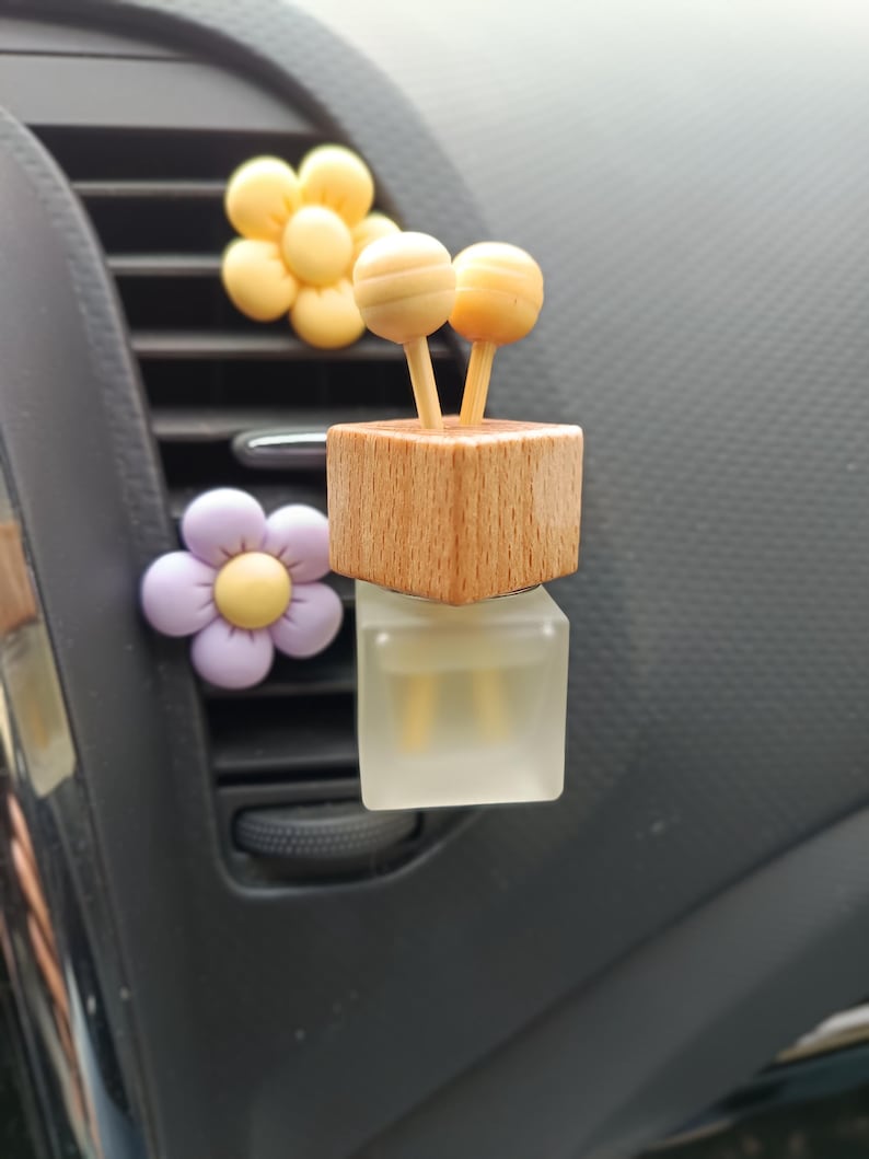 Op de afbeelding: Auto-geurverspreider met een houten blok en twee bolvormige toppen, bovenop een heldere, vierkante fles. Twee bloemvormige ventilatieclips in geel en paars zijn ook zichtbaar.