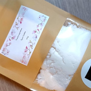 Könnte beinhalten: Eine Verpackung mit einem transparenten Fenster, das weißes Pulver zeigt. Ein Etikett mit floralen Akzenten liest sich "Poudre aspirateur Rose Vanille 100g". Ein runder Aufkleber mit einem schwarzen Quadrat ist vorhanden.