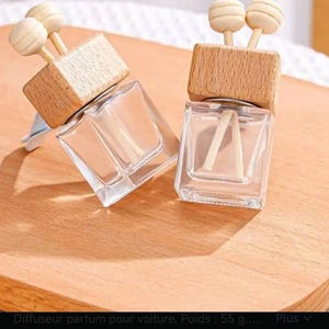 Op de afbeelding: Twee heldere glazen auto-geurverspreiders met houten tops en decoratieve houten accenten. Elk heeft een rechthoekige glazen fles met een houten dop en twee houten stokjes. De achtergrond is een lichtbruin oppervlak.