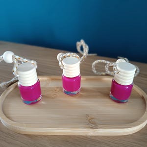 Op de afbeelding: Drie kleine, heldere glazen flesjes gevuld met felroze vloeistof, elk met een lichte houten dop. De flesjes staan op een licht houten dienblad, met een wit en goudkleurig touwtje eraan. De achtergrond is effen blauwgroen.