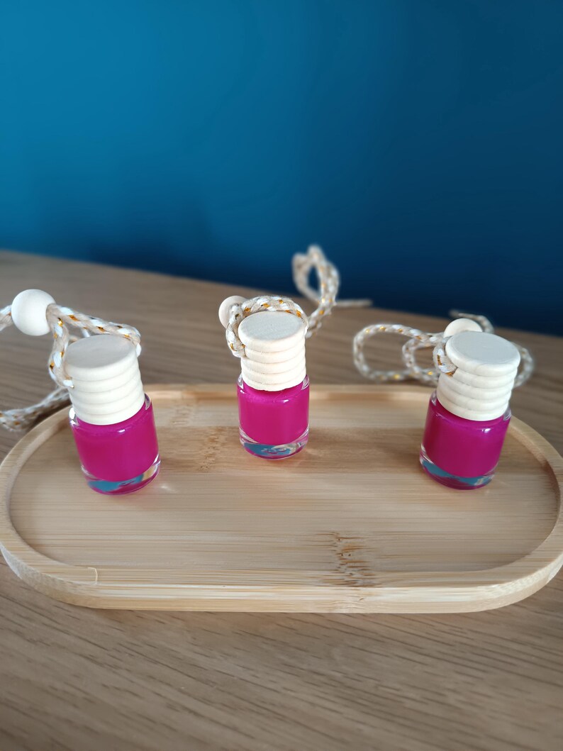 Op de afbeelding: Drie kleine, heldere glazen flesjes gevuld met felroze vloeistof, elk met een lichte houten dop. De flesjes staan op een licht houten dienblad, met een wit en goudkleurig touwtje eraan. De achtergrond is effen blauwgroen.