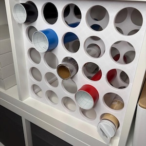 Könnte beinhalten: Ein weißes Aufbewahrungsregal mit kreisförmigen Aussparungen, das Rollen verschiedenfarbigen Geschenkpapieres enthält. Die Rollen sind schwarz, blau, rot und goldfarben. Das Regal steht auf einem weißen Regal.