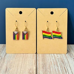 May include: Two pairs of gold-toned earrings displayed on brown cards. One pair features a rainbow flag with a purple triangle, the other pair has rainbow flag designs with the words "love wins". The earrings are set against a blue background.