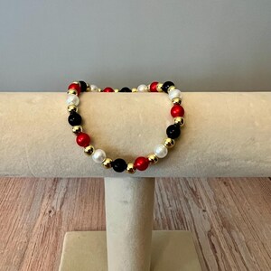 Peut inclure: Un bracelet de perles rouges, noires, blanches et dorées. Les perles sont disposées en cercle. Les perles sont rondes et espacées uniformément, avec un mélange de finitions mates et métalliques. Le bracelet est présenté sur un présentoir crème.