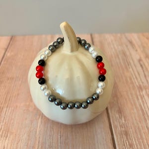 Könnte beinhalten: Ein Perlenarmband mit einer Mischung aus grauen, weißen, roten und schwarzen Perlen. Das Armband liegt auf einem weißen, kürbisförmigen Deko-Objekt vor einem hellen Holzgrund. Die Perlen haben einen Durchmesser von etwa 0,8 cm.