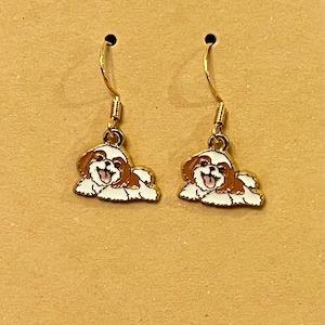 May include: A pair of gold-coloured drop earrings. Each earring has a small, enamelled charm shaped like a Shih Tzu dog. The dogs are white with brown markings and have their tongues sticking out.