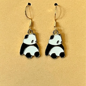 May include: A pair of gold-colored dangle earrings featuring panda bear designs. Each panda has a black and white color scheme with a gold-colored hook. The earrings are displayed against a light brown background.
