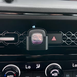 May include: Black car air vent clip-on air freshener with a purple and clear scent diffuser. The car's dashboard features climate control knobs, a hazard light button, and a diamond-patterned vent.