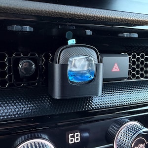 May include: A black car air freshener holder with a clear glass container filled with blue liquid. The holder is mounted on a car dashboard, near a hazard light button and air vent. The temperature display reads 68 degrees.