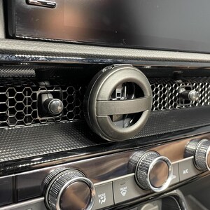 May include: A car dashboard close-up showing a black air vent with a circular air freshener. The dashboard has a honeycomb pattern and chrome dials. A digital display is visible in the background.