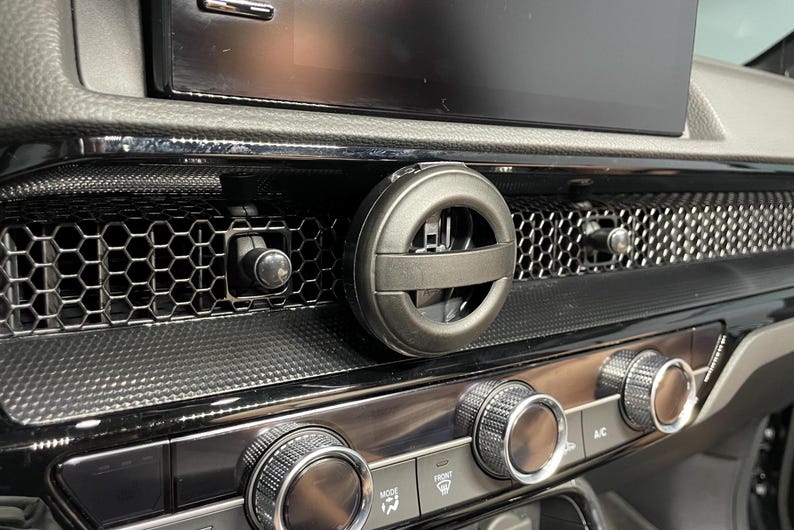 May include: A car dashboard close-up showing a black air vent with a circular air freshener. The dashboard has a honeycomb pattern and chrome dials. A digital display is visible in the background.