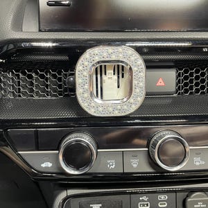 May include: A car air vent accessory with a silver frame and a square, crystal-studded design. The accessory is mounted on a black dashboard with various controls and a hazard light button.