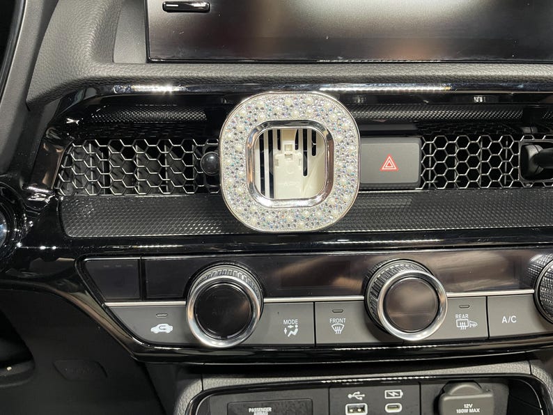 May include: A car air vent accessory with a silver frame and a square, crystal-studded design. The accessory is mounted on a black dashboard with various controls and a hazard light button.