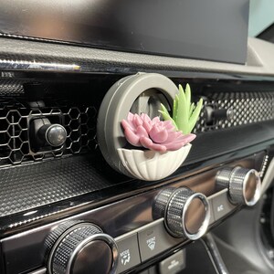 May include: A car air vent clip with a small, round, grey planter. The planter has a white, scalloped base and contains a pink and a green succulent. The clip is attached to a car dashboard.