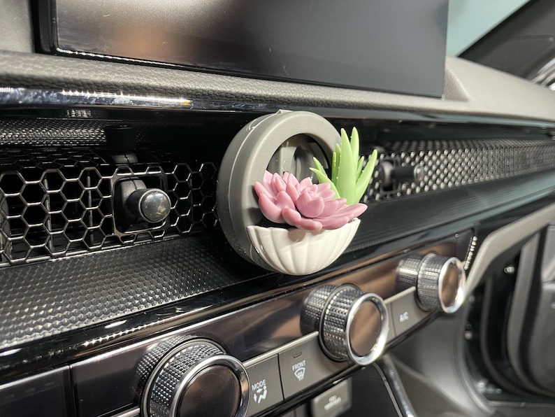 May include: A car air vent clip with a small, round, grey planter. The planter has a white, scalloped base and contains a pink and a green succulent. The clip is attached to a car dashboard.