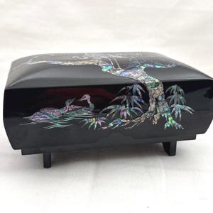 May include: A black lacquered box with iridescent mother-of-pearl inlay. The box features a tree and two ducks on the lid. The box is rectangular with short legs. The inlay has shades of green, blue, and purple.