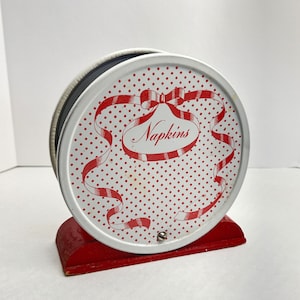 May include: A vintage napkin holder with a white, red polka-dot design. The holder has a red ribbon and the word "Napkins" in a decorative font. It sits on a red wooden base.