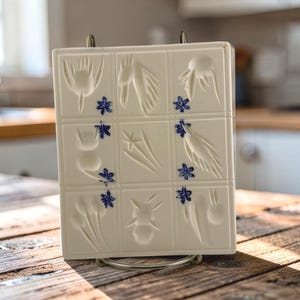 May include: A rectangular white ceramic mold featuring a grid of nine squares. Each square contains embossed botanical or abstract shapes, and small blue painted flower accents are positioned between the grid sections.