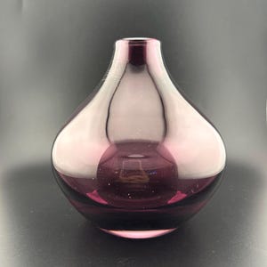 May include: A translucent purple glass vase with a rounded, bulbous body and a narrow, circular opening at the top. The glass features a smooth, polished surface that reflects light, creating a gradient of deep violet to lighter lavender tones.