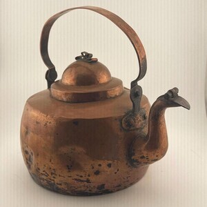 May include: A copper kettle with a rounded body, a curved spout, and a hinged lid featuring a small metal loop handle. The kettle has a tall, arched handle attached to the sides and displays a weathered, dark patina with visible surface wear.