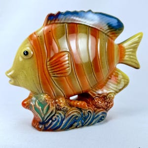 Puede incluir: Figura de pez de cerámica con una combinación de colores amarillo, naranja y azul. El pez tiene rayas verticales naranjas y está posado sobre una base con detalles de olas azules y verdes. Las aletas y la cola son amarillas.