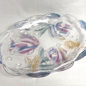 May include: Clear glass serving dish with a scalloped edge. The dish features embossed fish, seaweed, and seashell designs in shades of pink, blue, and yellow. The dish is oval-shaped.