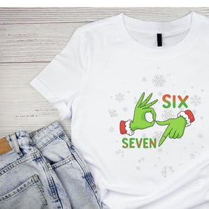 May include: White t-shirt with a green hand graphic forming the number six and seven with the text "SIX" and "SEVEN" in red and green. The hands are wearing red and white Santa hats. Snowflake designs are scattered around the graphic.