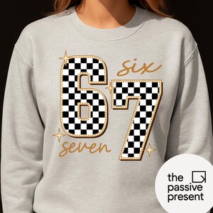 May include: A light gray sweatshirt with the number "67" in a black and white checkered pattern. The word "six" and "seven" are written in gold script. The sweatshirt has a round neck.
