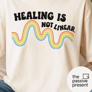 Puede incluir: Camiseta color crema con el texto negro "HEALING IS NOT LINEAR" sobre un diseño de arcoíris ondulado. La camiseta tiene cuello redondo y mangas cortas. El diseño incluye una onda de arcoíris en tonos rojo, naranja, amarillo, verde, azul y morado.
