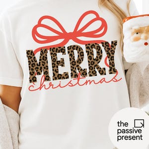 May include: White t-shirt with a red bow design above the words "MERRY" in leopard print and "christmas" in red script. The shirt is a festive holiday apparel item.