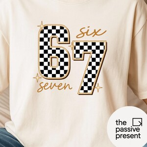 May include: Cream-colored t-shirt featuring the numbers "67" in a black and white checkered pattern, outlined in gold. The word "six" and "seven" are written in gold script. Gold starbursts accent the design. The text "the passive present" is in a white circle.