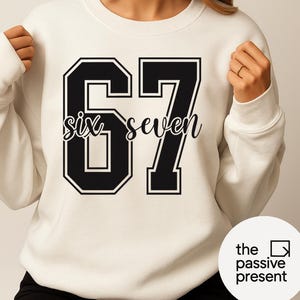 May include: Cream-colored sweatshirt with the number "67" in a black, collegiate-style font. The words "six seven" are written in cursive within the number. The sweatshirt has a crew neck and long sleeves. The brand logo is in the bottom right corner.
