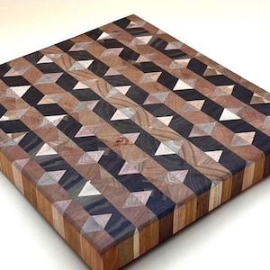 May include: A square wooden cutting board with a geometric pattern. The pattern is made up of various wood tones, including light brown, dark brown, black, and white. The board is approximately 12 inches square and has a smooth surface.
